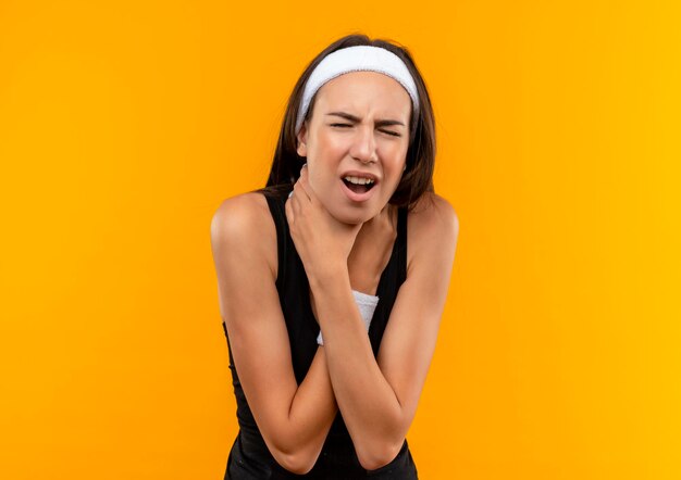 Fed up young pretty sporty girl wearing headband and wristband trying to choke herself on orange wall