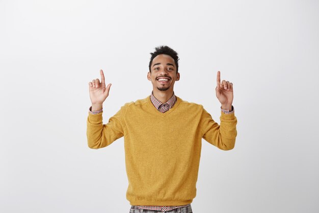 Чувствую себя победителем. Портрет довольного счастливого афро-американского парня в желтом пуловере, поднимающего указательные пальцы и указывающего вверх, довольного положительным результатом, торжествующего от хороших новостей