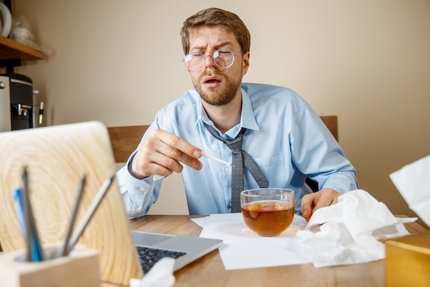 Feeling sick and tired. The man with cup of hot tea working in office, businessman caught cold, seasonal flu. Pandemic influenza, disease prevention, air conditioning in office cause sickness