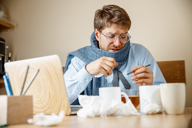 Feeling sick and tired. The man with cup of hot tea working in office, businessman caught cold, seasonal flu. Pandemic influenza, disease prevention, air conditioning in office cause sickness