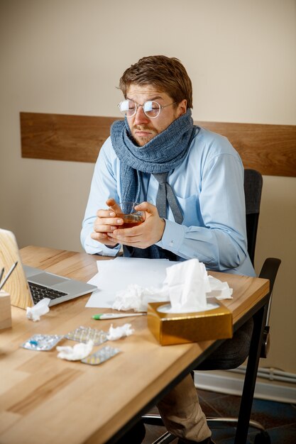 Feeling sick and tired. The man with cup of hot tea working in office, businessman caught cold, seasonal flu. Pandemic influenza, disease prevention, air conditioning in office cause sickness