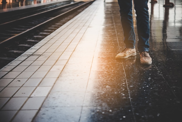 鉄道駅のプラットフォームで列車を待っているジーンズを着た若者の足。