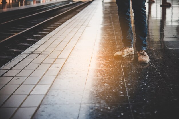 鉄道駅のプラットフォームで列車を待っているジーンズを着た若者の足。