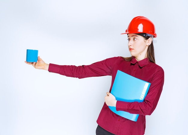 Female architect in red helmet documents in hand holding coffee. 