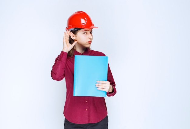 Female architect in red helmet holding blue folder and posing to camera. 