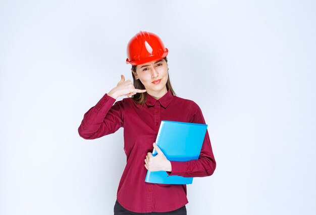 파란색 폴더를 들고 카메라에 포즈를 취하는 빨간 헬멧에 여성 건축가.