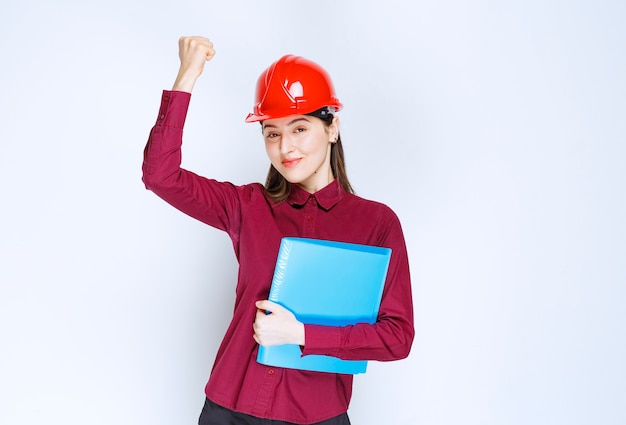 Female architect in red helmet holding blue folder with documents. 