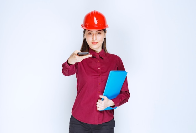 Female architect in red helmet holding clipboard and giving away cellphone. 