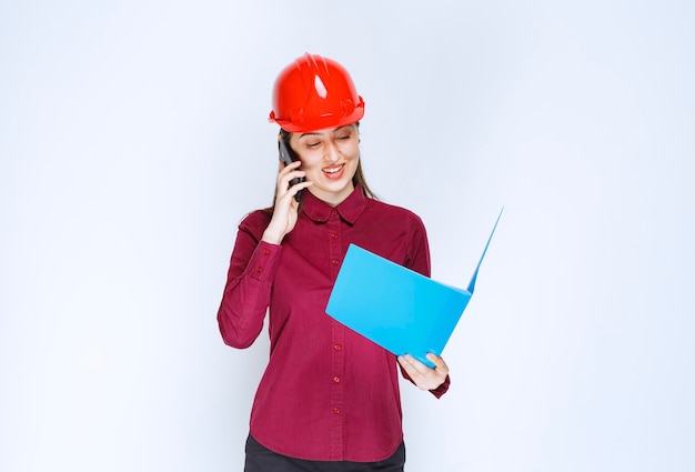 Female architect in red helmet holding clipboard and talking via cellphone. 