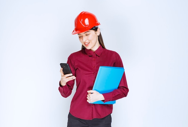 Женский архитектор в красном шлеме сзажимом для бумаги и разговором по мобильному телефону.