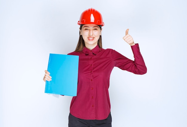 Female architect in red helmet with blue folder getting standing and posing. 