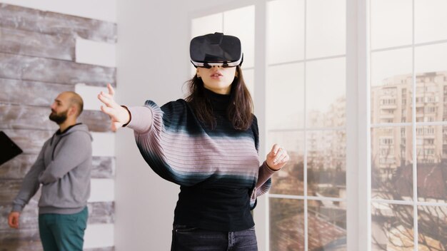 Female architect using virtual reality goggles in empty apartment and real estate agent talking with client in the background.