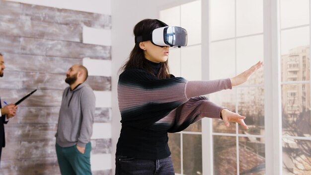 Female architect using virtual reality goggles in empty apartment and real estate agent talking with client in the background.