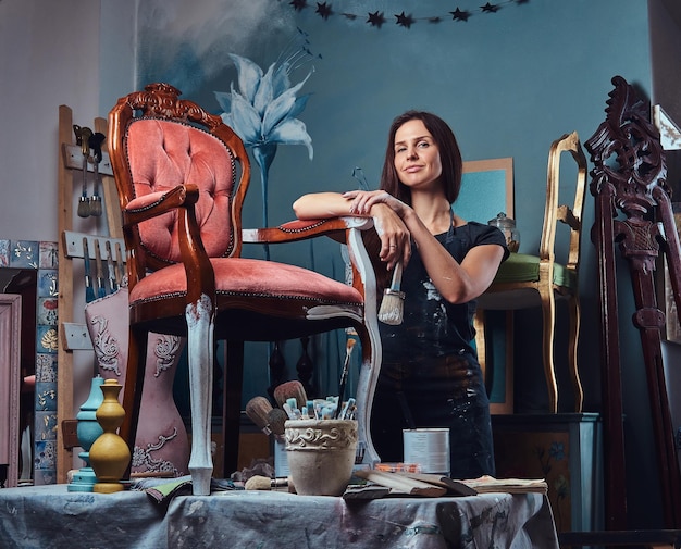 Female artist in apron holds paintbrush and leaning on a vintage chair in workshop.