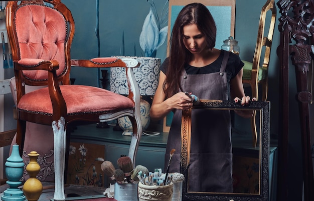 Female artist in apron painting vintage picture frame with paintbrush in workshop.