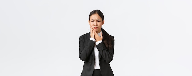 仕事で歯痛を抱えている黒いスーツの女性アジア人オフィスマネージャー歯の痛みを感じているので頬に手を握っている問題を抱えた実業家は白い背景に立っている予約医師が必要です