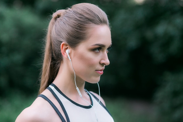 女性の競技者はイヤホンを着用しています。屋外で運動中に音楽を聴く女性。