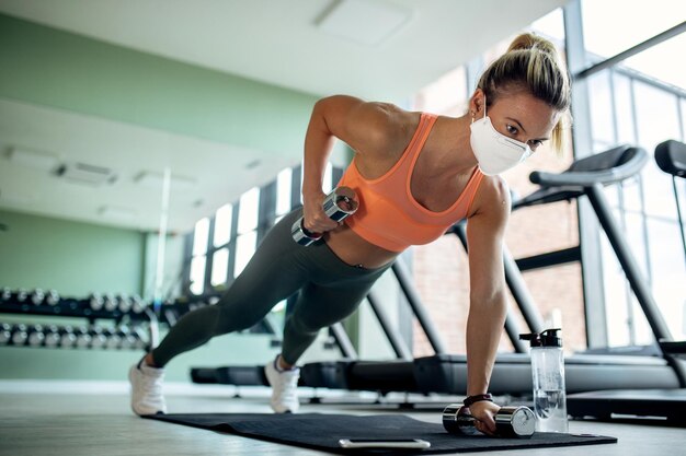 Female athlete with protective face mask doing plank exercise with hand weights in a gym