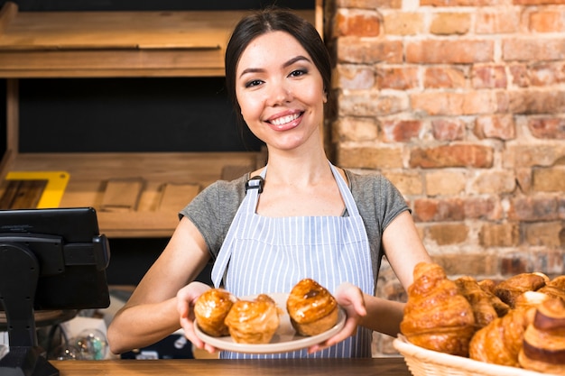 빵집 가게 카운터에서 접시에 구운 달콤한 퍼프 페이스 트리를 보여주는 여성 베이커