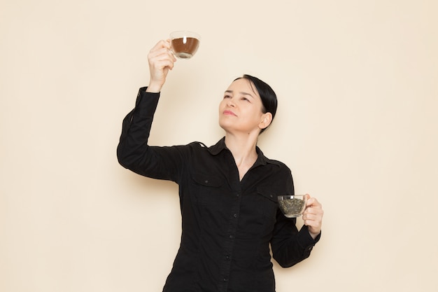 female barista in black shirt trousers holding cups with dried tea and coffee on the white wall