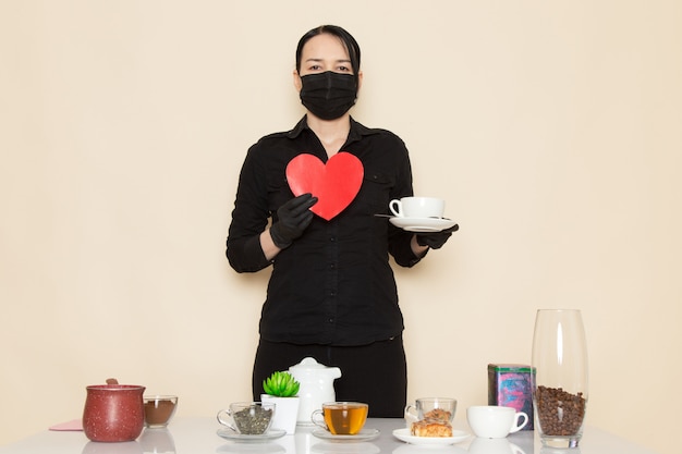 female barista in black shirt trousers with coffee brown dried tea equipment ingredients in black sterile mask on the white wall