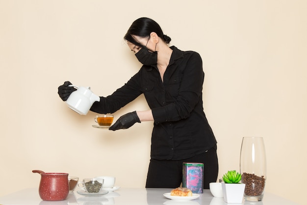 Female barista in black shirt trousers with coffee brown dried tea equipment ingredients in black sterile mask on the white wall