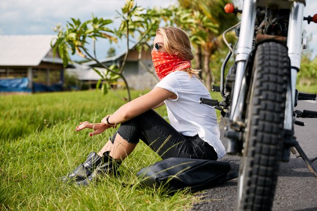 Female biker sitting on grass next to motorbike