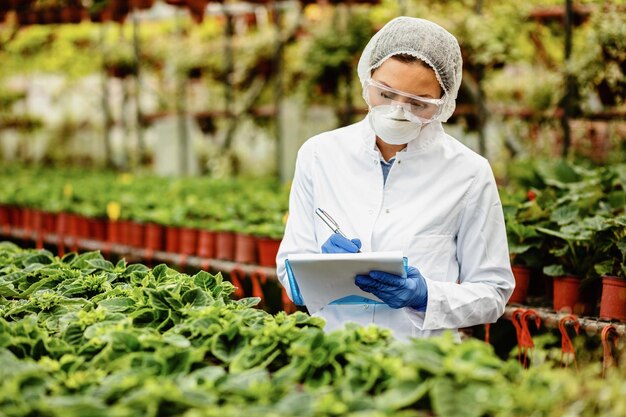 Female biotechnologist inspecting potted plants in plant nursery and writing notes into clipboard