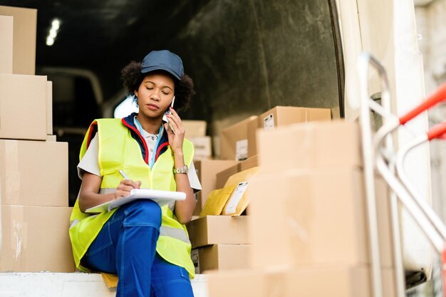 Female black courier sitting in delivery van and communicating on mobile phone while writing notes in clipboard