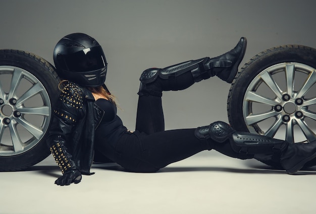 Female in black motrcycle helmet and leather clothes lying between two car wheels.