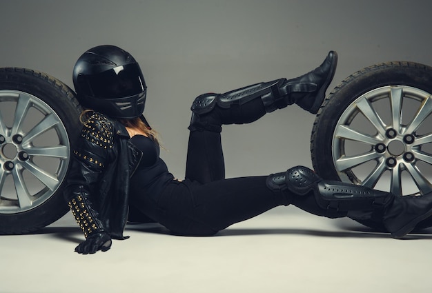 2つの車の車輪の間に横たわっている黒いオートバイのヘルメットと革の服を着た女性。