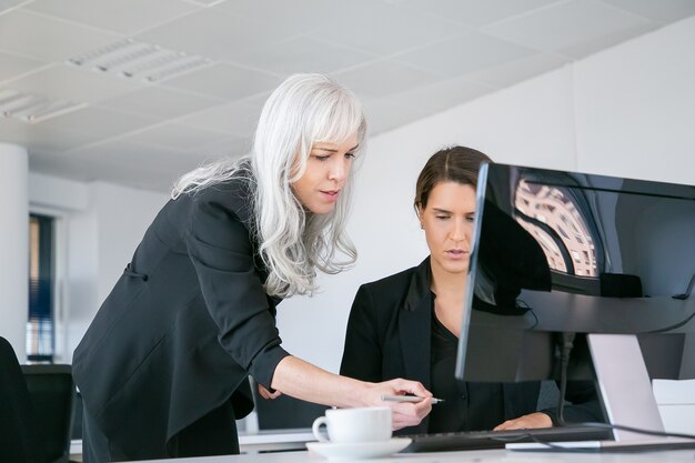 Female boss affixing signature on managers report. Businesswomen sitting and standing at workplace with monitor and coffee cup. Business communication concept