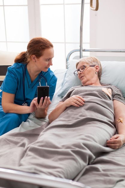 무료 사진 요양원의 여성 간병인은 침대에 앉아 휴대폰을 사용하는 노부인과 이야기를 나눴습니다.