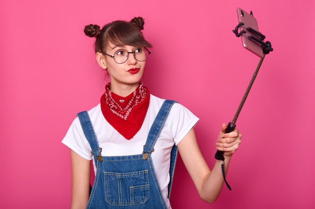 Female in casual white t shirt, overalls, red bandana on neck and round glasses. Adorable teenager keeps lips rounded