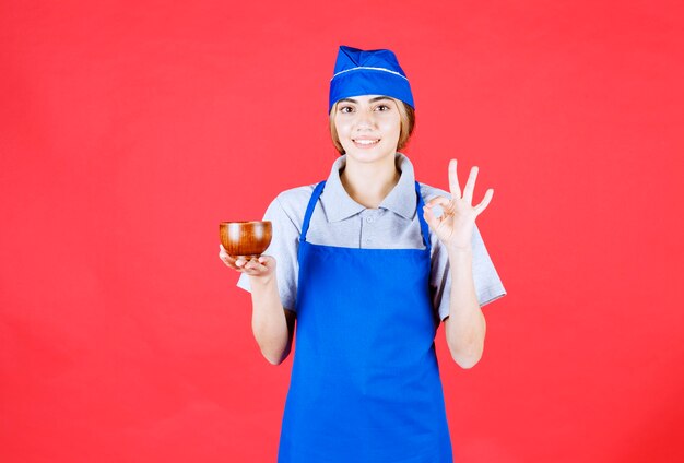 파란색 앞치마를 입은 여성 요리사는 중국 구리 찻잔을 들고 맛에 대해 긍정적인 느낌을 받습니다.