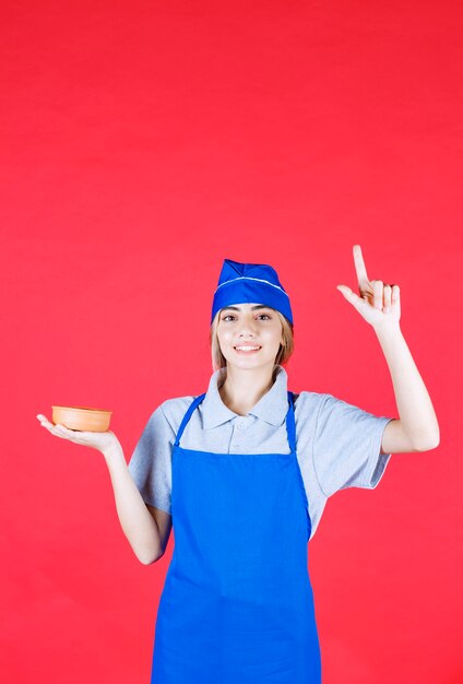 Женщина-повар в синем фартуке держит чашку с лапшой и выглядит сбитой с толку и думает о том, как сделать ее вкуснее