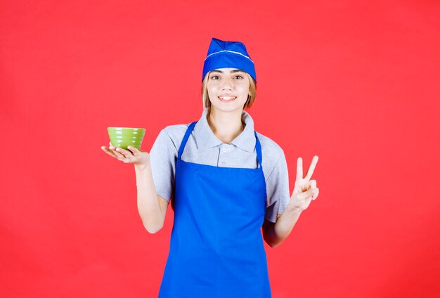 Female chef in blue apron holding a noodle cup and showing satisfaction hand sign