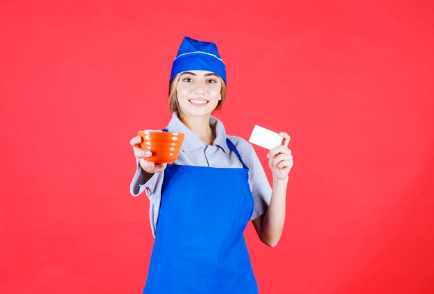 Шеф-повар в синем фартуке держит оранжевую керамическую чашку с лапшой и представляет свою визитную карточку