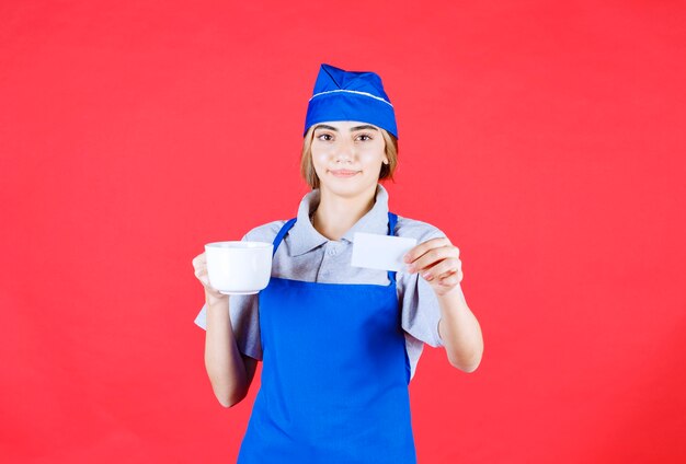 Free photo female chef in blue apron holding a white ceramic noodle cup and presenting her business card