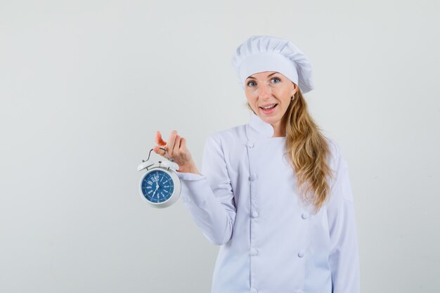 Female chef holding alarm clock in white uniform and looking cheery 