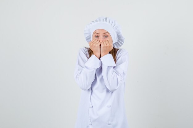 Female chef holding fists on mouth in white uniform and looking scared. front view.