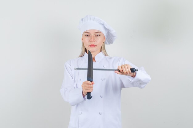 Женщина-повар держит нож и точилку в белой форме и выглядит серьезно