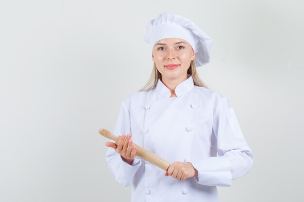 Female chef holding rolling pin in white uniform and looking cheery.