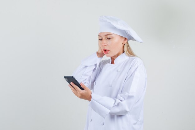 Женский шеф-повар держит смартфон с рукой на голове в белой форме и выглядит удивленным.