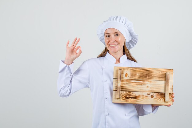 Cuoco unico femminile che tiene vassoio di legno con il segno giusto in uniforme bianca e che sembra allegro