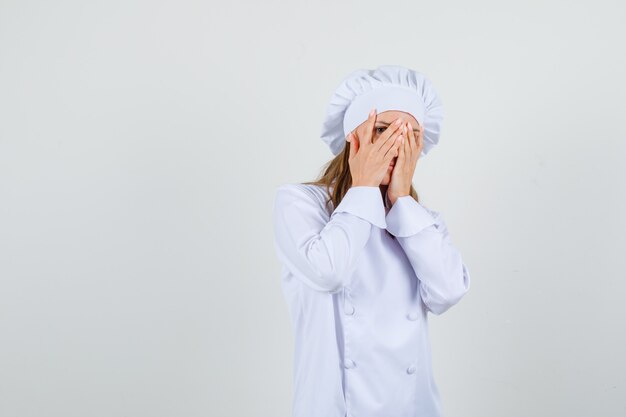 Female chef peeking through fingers in white uniform and looking positive. front view.