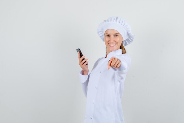 Female chef pointing finger at camera with smartphone in white uniform and looking cheerful. front view.