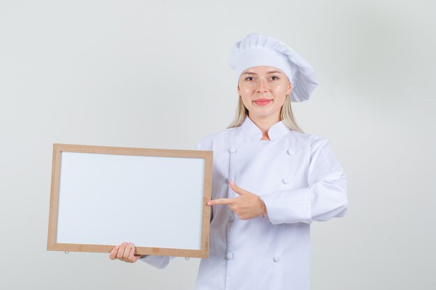 Female chef pointing finger at white board in white uniform and looking cheery.