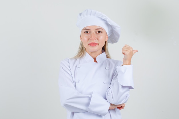 Female chef pointing thumb to side in white uniform and looking cheerful 