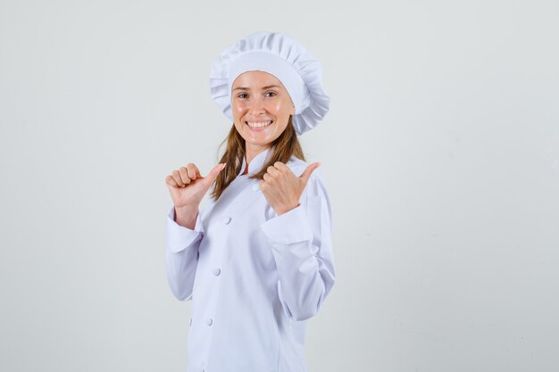 Female chef pointing thumbs to side in white uniform and looking cheerful .